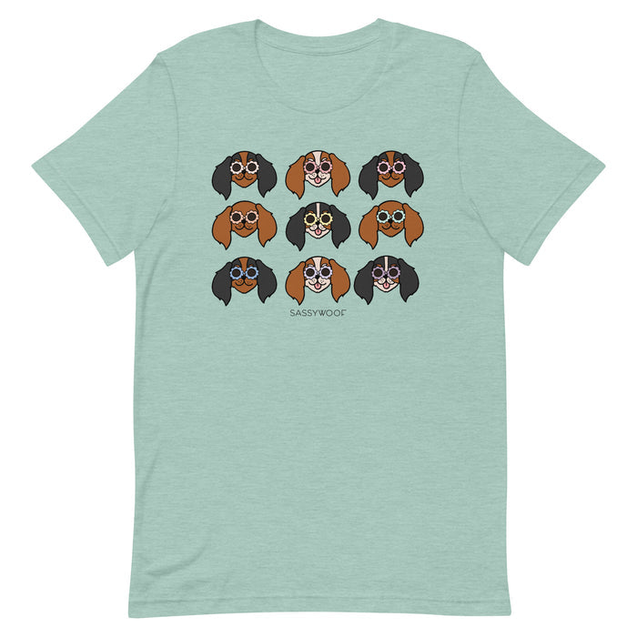 The Sassy Woof Cavalier Tee is a light green unisex 100% cotton shirt with nine unique cartoon dogs in glasses and "SASSY WOOF" printed below—the perfect playful touch for any outfit.