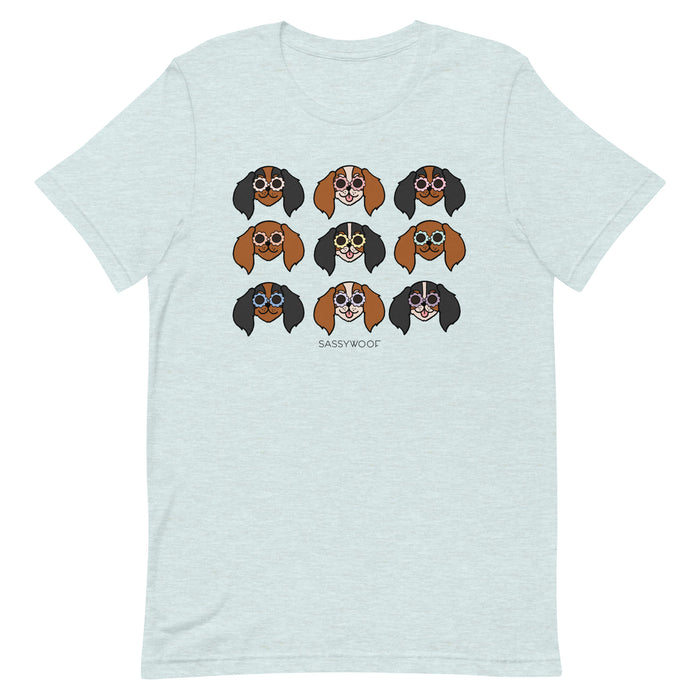 The Cavalier Tee by Sassy Woof is a light gray unisex t-shirt with nine illustrated dogs in sunglasses, arranged in three rows, showing various brown, black, and white fur patterns with "SASSY WOOF" text below.