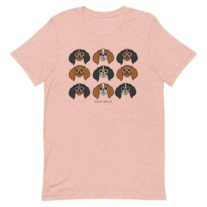 The Sassy Woof Cavalier Tee is a light pink unisex 100% cotton t-shirt with a grid of nine cartoon dogs in colorful sunglasses and "SASSYWOOF" printed beneath the playful design.