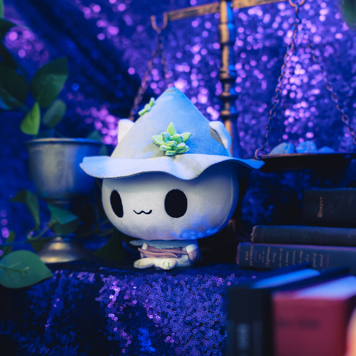 Cloudheads: 5" Mystic Moon (Fern). A cute white plush cat with large black eyes, a small wavy smile, wearing a gray pointed hat with green leaf accents and a brown skirt, shown with a sparkly purple backdrop as well as a leafy green plant and a pile of books.