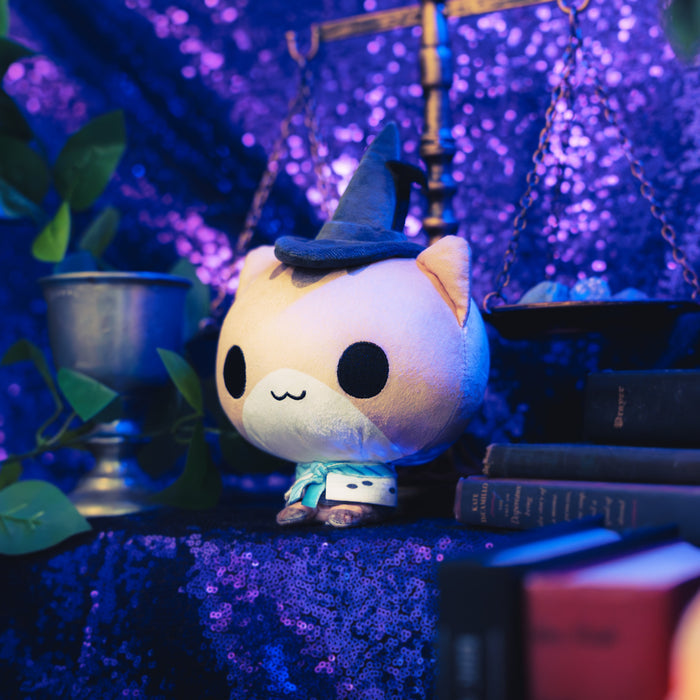 Cloudheads: 5" Mystic Moon (Melody) -  A plush toy of a cute cat with a black witch hat and blue scarf,  shown with a sparkly purple backdrop as well as a leafy green plant and a pile of books.