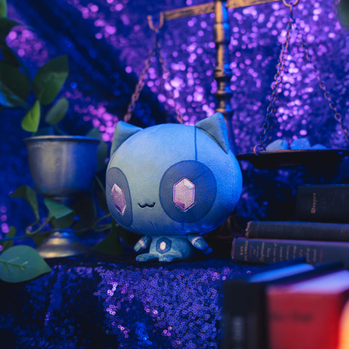 Cloudheads: 5" Mystic Moon (Quartz) - A blue cat plush toy with large dark blue eyes with pink gem-shaped crystals in the center, and a small black stitched smile, shown with a sparkly purple backdrop as well as a leafy green plant, a goblet and a pile of books.