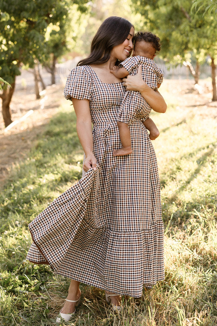 A woman wearing the Ivy City Co Connie Short Sleeve Dress in Chestnut Gingham holds a baby dressed in matching clothes, standing outdoors on grass with trees behind them as sunlight filters through the scene.