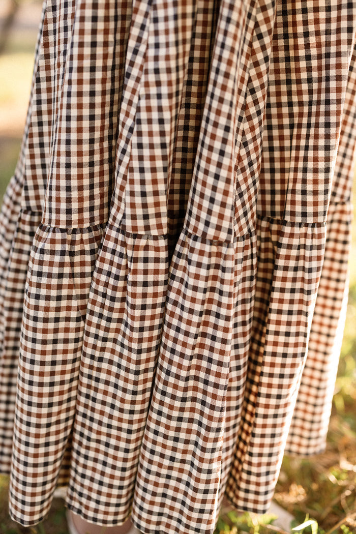The Connie Short Sleeve Dress in Chestnut Gingham by Ivy City Co features brown and white gingham-patterned fabric with gathered, pleated sections, elegantly displayed outdoors in natural sunlight.