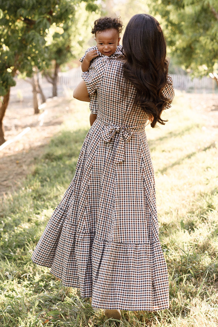 A woman wearing the Ivy City Co Connie Short Sleeve Dress in Chestnut Gingham holds a smiling baby as they walk through a sunlit orchard, both seen from behind amid green trees and soft, natural light.