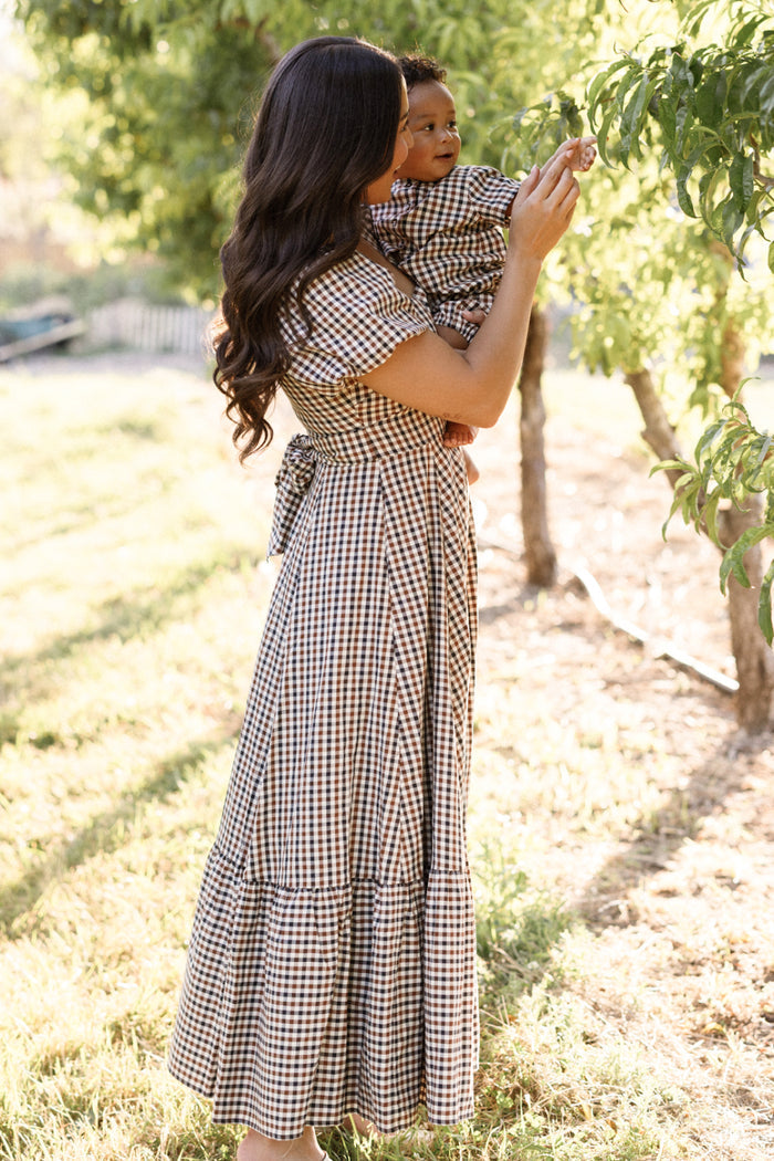 A woman wearing the Ivy City Co Connie Short Sleeve Dress in Chestnut Gingham holds a baby dressed in a matching outfit while standing outdoors near green trees on a sunny day.