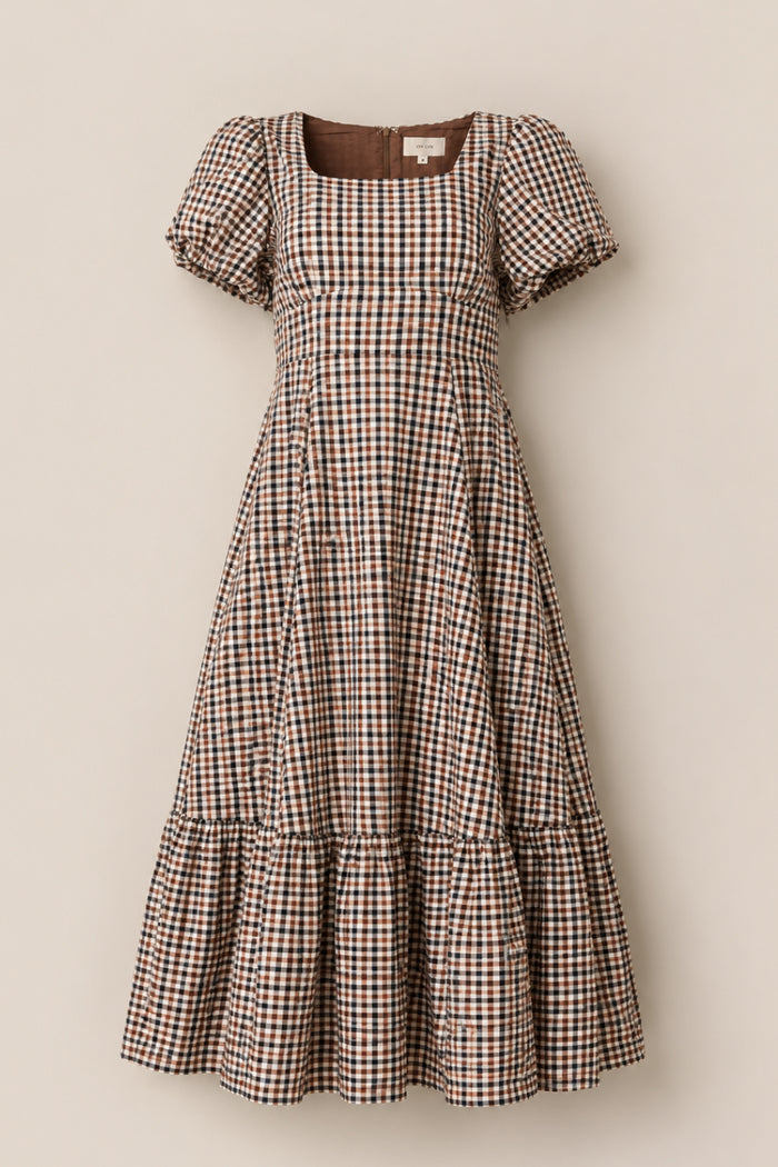 The Connie Short Sleeve Dress in Chestnut Gingham by Ivy City Co features a knee-length, flared skirt with short puff sleeves and a ruffled hem. Designed with a brown, black, and white gingham check pattern, this dress showcases a square neckline and fitted bodice for a classic silhouette.