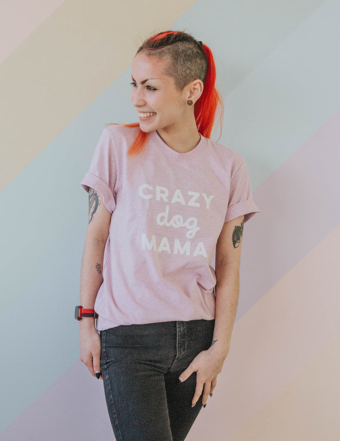 A smiling red-haired person with a side shave wears the Sassy Woof Crazy Dog Mama Tee and black jeans, standing against a pastel-striped background.