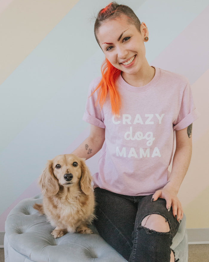 A smiling person with bright orange hair in a ponytail sits on a stool beside a small, long-haired tan dog, wearing the Sassy Woof Crazy Dog Mama Tee and ripped black jeans against a pastel striped background.