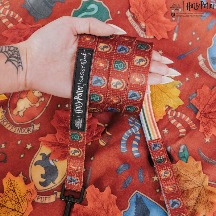 A hand with long, pale nails holds a Sassy Woof Dog Leash - Harry Potter™ featuring house crests. The background is red fabric adorned with Hogwarts house emblems, autumn leaves, candy, and matching pet accessories.