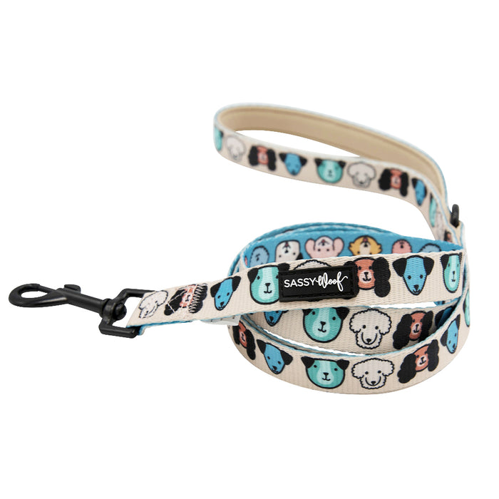 The Sassy Woof "Mixed With Love" dog leash features a cream design with colorful cartoon dog faces, a comfy neoprene handle, sturdy black metal clip, and signature Sassy Woof label.