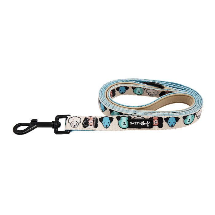 The “Mixed With Love” dog leash by Sassy Woof features cartoon dog faces in blue, black, and pink on cream, a soft neoprene handle, a black metal clip, and a visible Sassy Woof brand label.