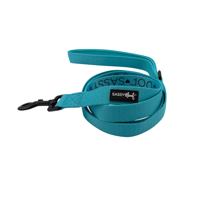 The Sassy Woof Dog Leash in Neon Blue features a black metal clasp, a black label with the brand name, and a soft neoprene handle. The leash is partially coiled on a white background.