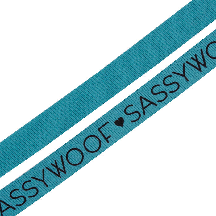 Two neon blue fabric straps are shown diagonally. One strap, part of the Dog Leash - Neon Blue by Sassy Woof, features “SASSY WOOF” repeatedly printed in black with a heart symbol between the words.