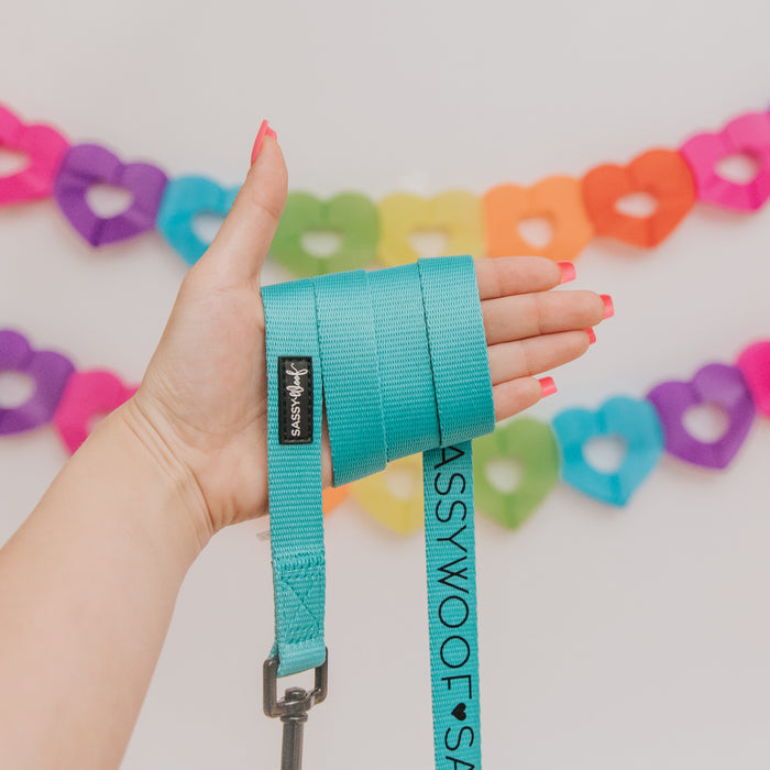 A hand with pink-painted nails holds the Sassy Woof Dog Leash - Neon Blue, featuring a neoprene handle, against a backdrop of vibrant heart-shaped paper garlands.