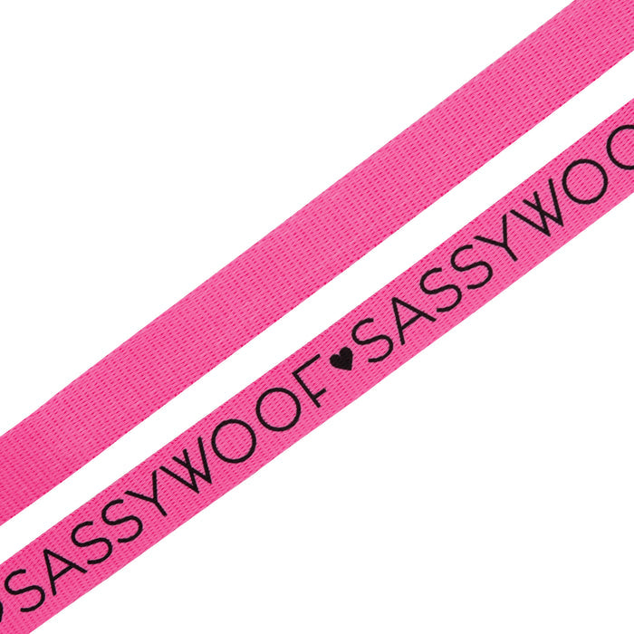 Two neon pink fabric straps from Sassy Woof, each printed with “SASSY WOOF” in black and separated by a black heart—perfect for pairing with your favorite adjustable dog harness or other neon accessories.