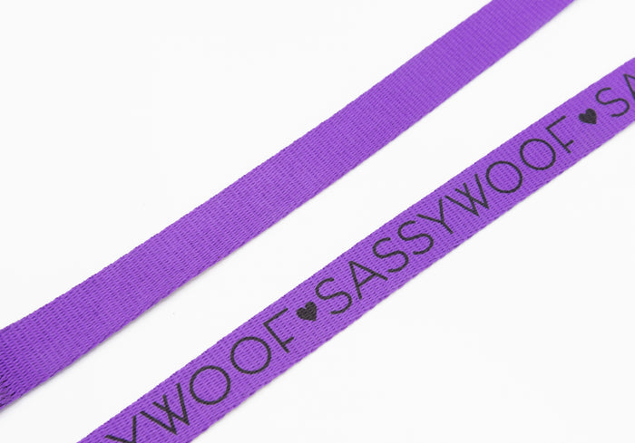 A close-up of two neon purple Sassy Woof dog leashes on a white background. One leash features “SASSY WOOF” and heart symbols printed in black.