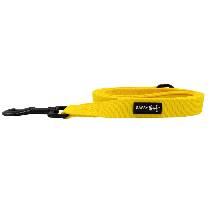 The Sassy Woof Dog Leash - Neon Yellow features a bright yellow color, black metal clasp, and a Sassy Woof fabric label. The leash is shown coiled neatly against a white background.