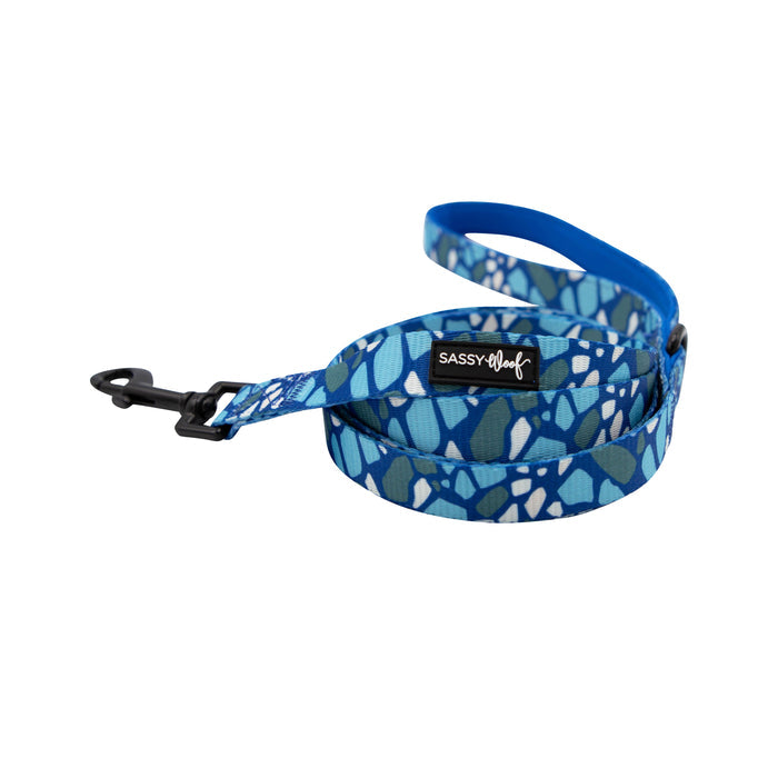 The Sassy Woof Dog Leash - Santorini features a blue abstract pattern, soft neoprene handle, and black metal clip. Shown coiled on a white background. Pair it with the matching Santorini harness for a coordinated look.
