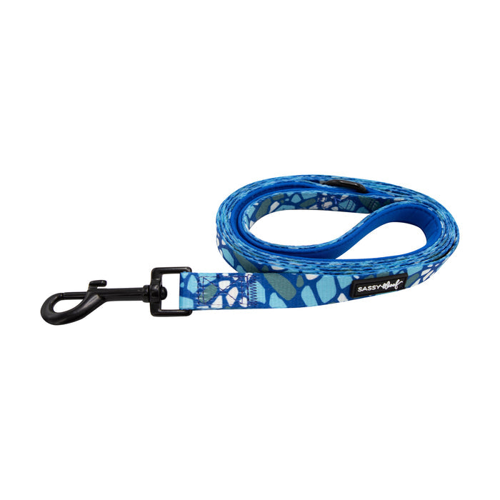The Sassy Woof Dog Leash - Santorini features a blue pattern, neoprene handle, and black metal clip. Labeled with the brand name, it’s shown coiled on white and pairs perfectly with the matching harness.