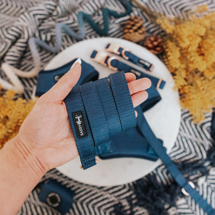 A hand holds the Sassy Woof Dog Leash - Twilight, a navy blue leash with rose gold hardware and branding. Matching pet accessories and yellow flowers rest on a patterned fabric background.