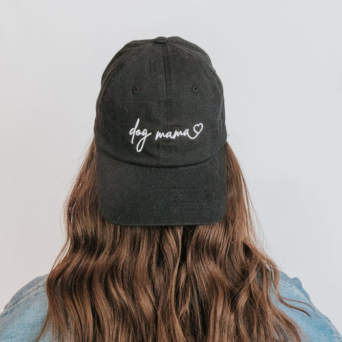 A person with long wavy brown hair is seen from behind wearing the Sassy Woof Dog Mama Hat - Black, featuring white "Dog Mama" embroidery and a small heart, adjustable strap, set against a light background.