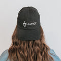 A person with long wavy brown hair is seen from behind wearing the Sassy Woof Dog Mama Hat - Black, featuring white "Dog Mama" embroidery and a small heart, adjustable strap, set against a light background.