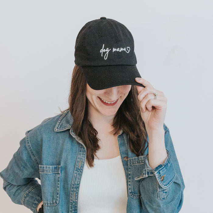 A woman in a denim shirt and white top smiles while holding the brim of her Sassy Woof Dog Mama Hat – Black, an embroidered cap with a heart detail and adjustable strap for the perfect fit.