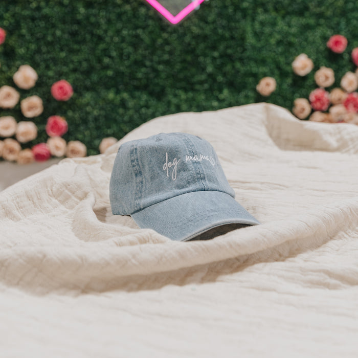 The Dog Mama Hat - Denim by Sassy Woof, a light blue cap with "dog mama" embroidered on the front, sits on a beige blanket among green foliage and pink and cream flowers.