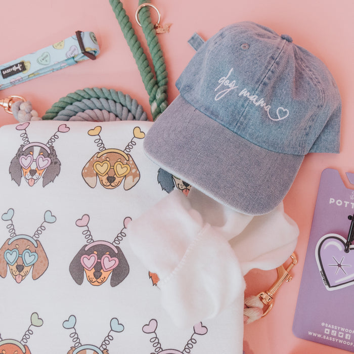 A flat lay of dog-themed items on a pink background features the Sassy Woof Dog Mama Hat in denim, a rope leash, a tote with illustrated dog faces, a purple tag, and a white scrunchie.