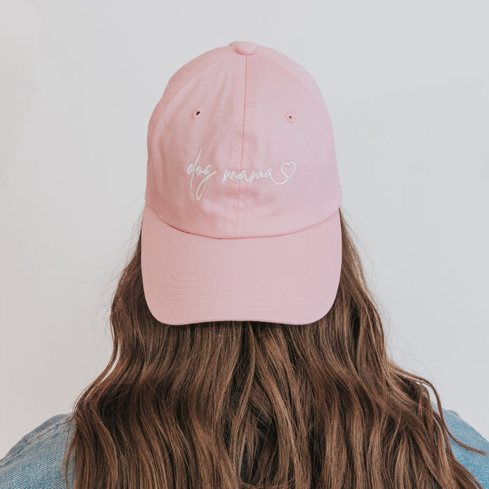 A person with long brown hair faces away from the camera, wearing the Sassy Woof Dog Mama Hat - Pink, a light pink adjustable baseball cap with "dog mama" and a heart embroidered in white thread.