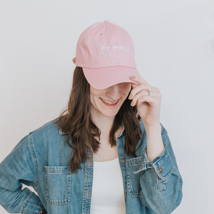 A woman with long brown hair in a denim jacket and white top smiles while touching the brim of her Sassy Woof Dog Mama Hat - Pink, an adjustable cap perfect for every proud pup parent.