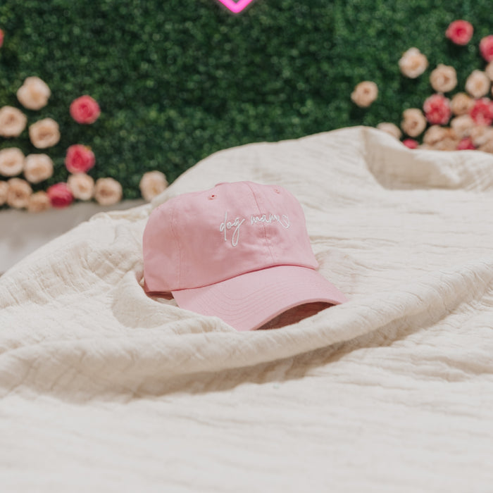 The Sassy Woof Dog Mama Hat - Pink with white cursive embroidery sits on a cream textured blanket, while a green hedge with pink and cream flowers enhances the charm of this adjustable dog mom hat scene.