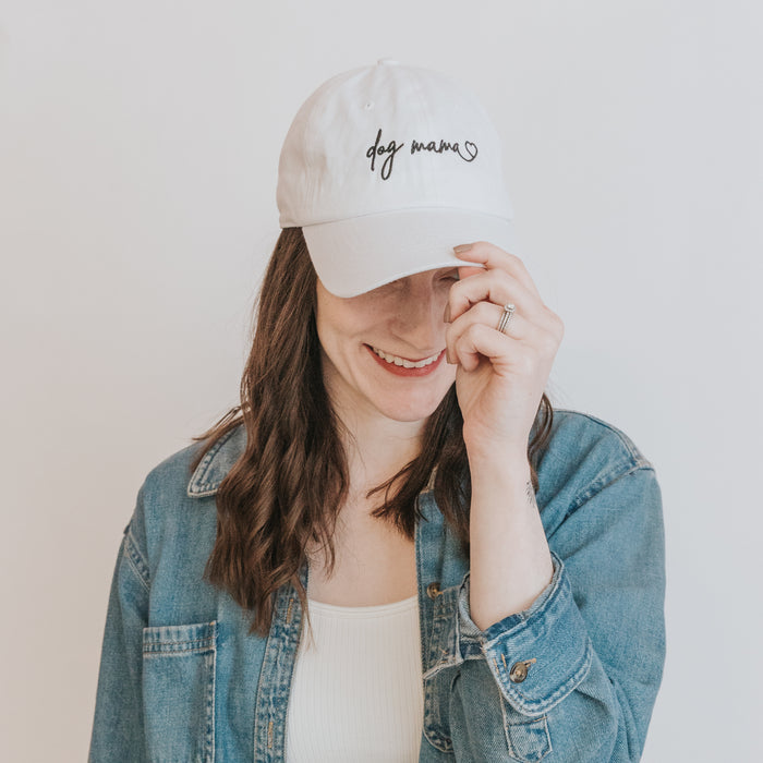 A woman in a denim jacket smiles, holding the brim of her Sassy Woof Dog Mama Hat - White. The adjustable hat features black script and pairs perfectly with her long brown hair and casual outfit against a plain background.