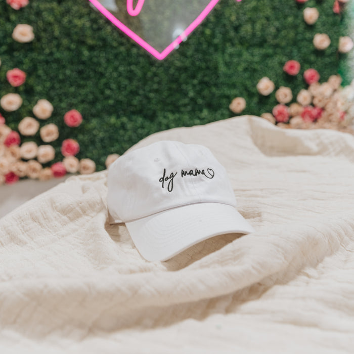 The Sassy Woof Dog Mama Hat in white features "dog mama" and a small heart embroidered on the front, shown on a cream blanket with a green and pink floral background.