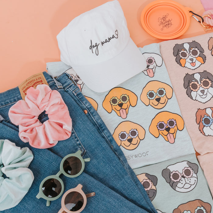 Flat lay of dog-themed t-shirts, the Sassy Woof Dog Mama Hat in white, blue jeans, colorful scrunchies, green and pink sunglasses, and an orange Save them All pet bowl on a peach background.