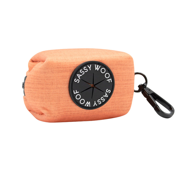 The Sassy Woof Dog Waste Bag Holder - Apple Cider features orange fabric, a black circular logo, and a black clip for attaching to a leash or bag—ideal for pairing with other dog walk accessories.