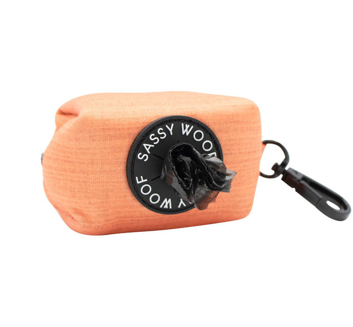 The Sassy Woof Dog Waste Bag Holder - Apple Cider is an orange fabric holder with a black clip and Sassy Woof label. It dispenses black bags easily and matches perfectly with the coordinating Apple Cider harness.