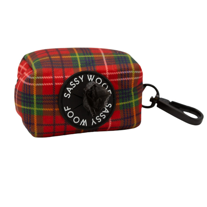 The Sassy Woof "Deck the Paws" dog waste bag holder features red and green tartan plaid flannel, a black clip, and a circular Sassy Woof label around the bag opening.