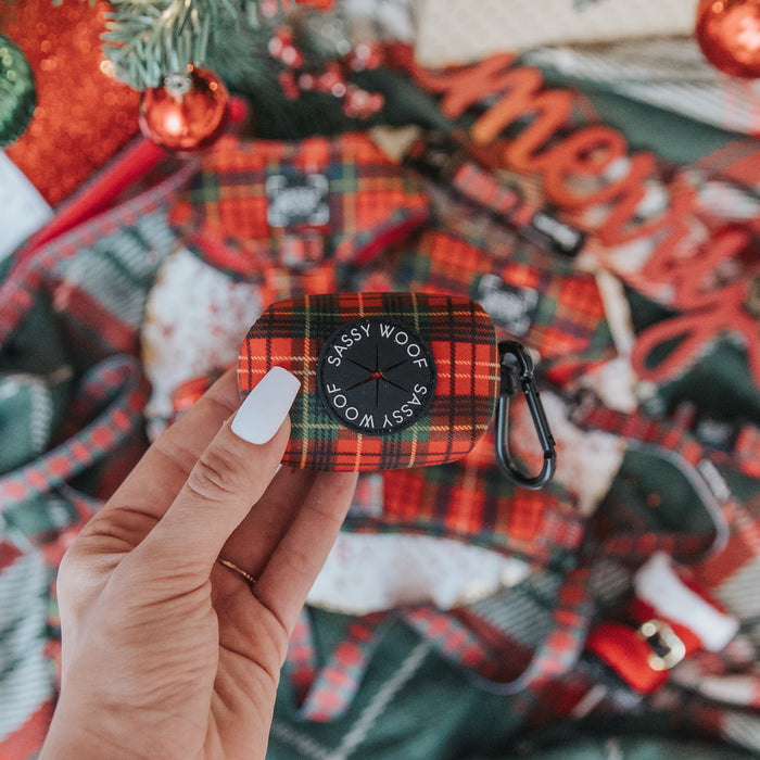 A hand with white-painted nails holds the Sassy Woof Dog Waste Bag Holder - Deck the Paws, with Christmas decorations and plaid fabric blurred festively in the background.