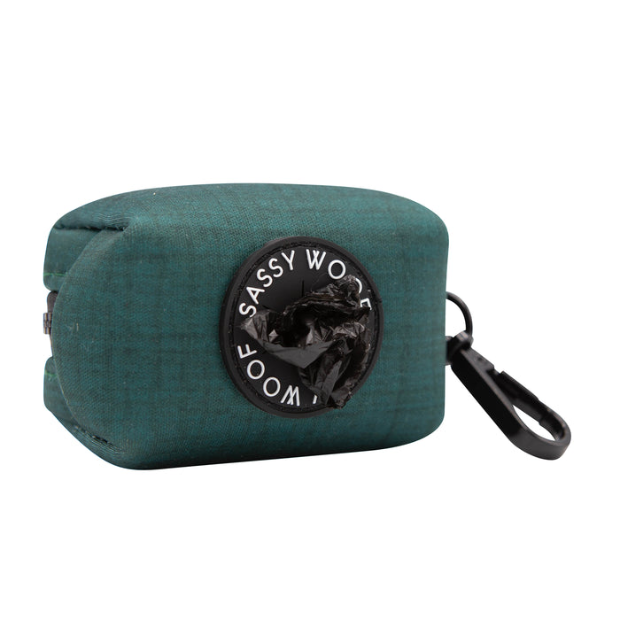 The Sassy Woof Dog Waste Bag Holder - Forest features teal neoprene, a black clip, circular dispenser with black bags, and branded detailing around the opening.