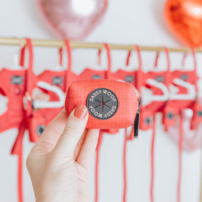 A hand holds the Sassy Woof Dog Waste Bag Holder - Merlot in focus, with red harnesses and heart-shaped balloons blurred behind—a chic neoprene dog accessory perfect for walks.