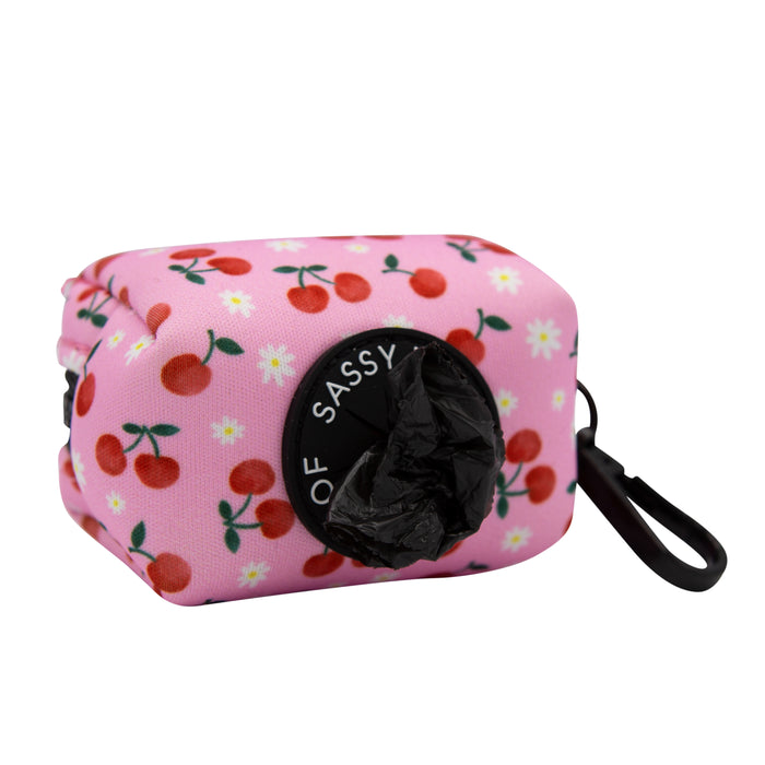 The Sassy Woof Dog Waste Bag Holder - Mon Chérry is pink with cherry and flower designs, featuring a black circular opening with a partially pulled bag and a black strap for easy attachment.