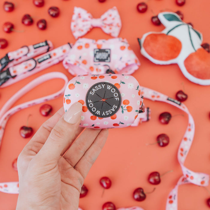 A hand holds the Sassy Woof Dog Waste Bag Holder - Mon Chérry, featuring a cherry pattern. Matching cherry-themed Sassy Woof leash, harness, and toy are scattered on an orange surface with fresh cherries.