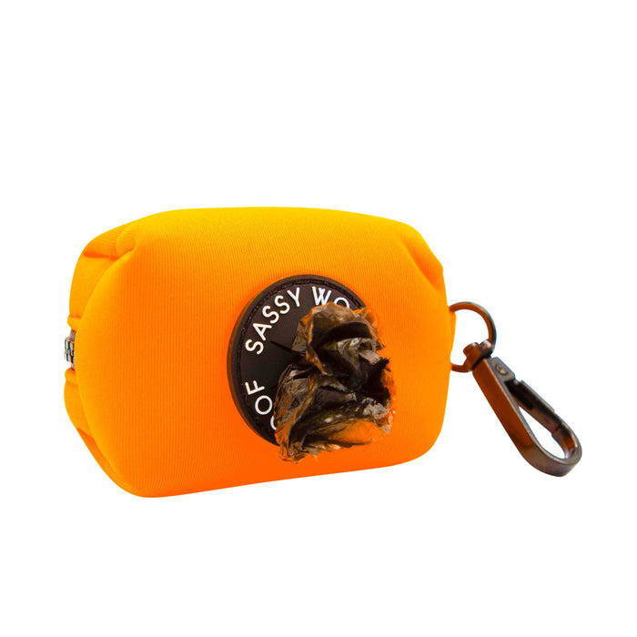 The Sassy Woof Dog Waste Bag Holder in Neon Orange features a circular dispenser, side brown clip, and partially visible black bag. Perfect to pair with your matching harness set. Background is white.