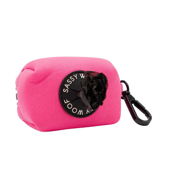 The Sassy Woof Dog Waste Bag Holder - Neon Pink features bright neoprene, a round black label, and a black carabiner clip, dispensing bags from the side—perfect to pair with your matching dog harness set.