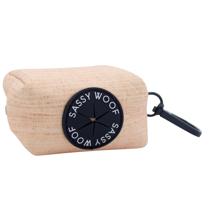 The Dog Waste Bag Holder - Pinot by Sassy Woof is a beige holder featuring a black circular Sassy Woof logo and a black clip for easy carrying—ideal to pair with your adjustable harness or any minimalist dog accessories.