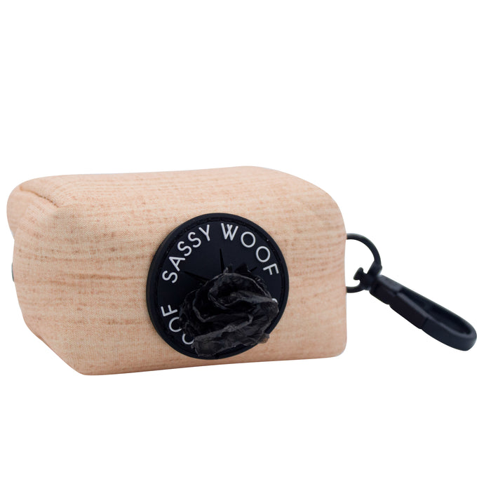 The Sassy Woof Dog Waste Bag Holder - Pinot features a beige design with a black circular dispenser, branded Sassy Woof, and a black clip for easy attachment. A partially pulled-out black waste bag offers added convenience for walks.
