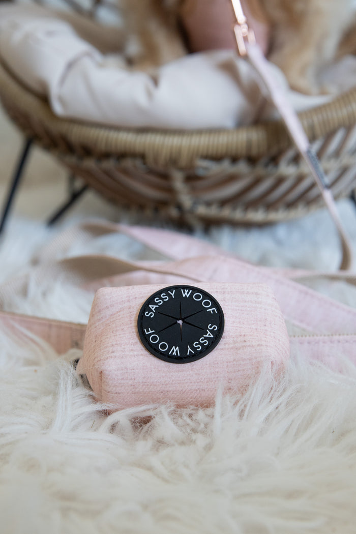 A pink Sassy Woof bag with a round black logo rests on a fluffy rug, paired with the Sassy Woof Dog Waste Bag Holder - Pinot, an adjustable harness, and a pink leash near a wicker pet bed in the background.