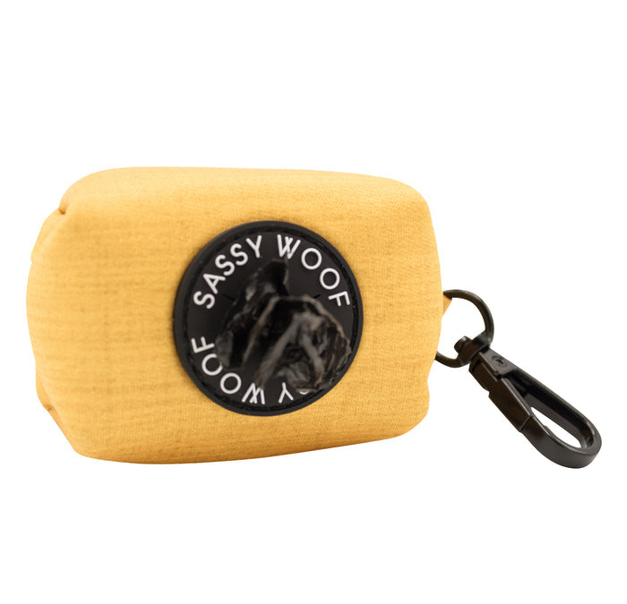 The Sassy Woof Dog Waste Bag Holder - Sunflower Fields is a small, rectangular yellow neoprene holder with a black circular Sassy Woof patch and plastic clip—an essential dog walking accessory for easy waste bag access.